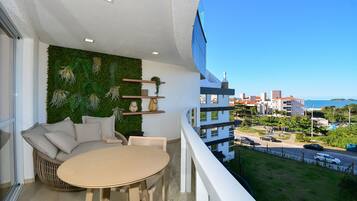 A balcony with a round table and a wicker sofa, a green wall with plants, and a view of buildings and the sea.