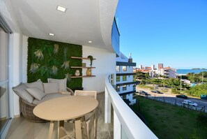 A balcony with a round table and a wicker sofa, a green wall with plants, and a view of buildings and the sea. - Gaivotas Golden Luxo a 200m praia-NIN012 (Florianópolis)