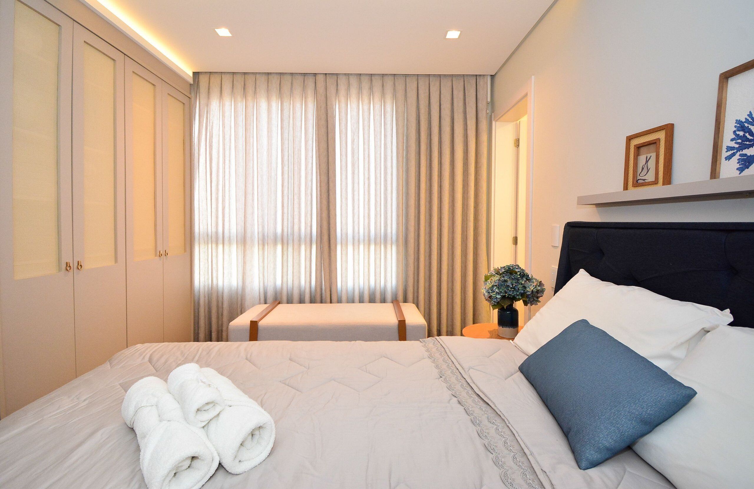 A hotel room with a bed, a bench, a nightstand with a vase, and a wall-mounted shelf with a picture.
