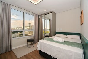 A hotel room with a large window, a bed with white linens, a green headboard, and a small stool. - Gaivotas Golden Luxo a 200m praia-NIN012 (Florianópolis)