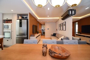 A modern kitchen with built-in appliances, a dining area with a wooden table and a decorative sculpture, and a living area with a sofa and television. - Gaivotas Golden Luxo a 200m praia-NIN012 (Florianópolis)