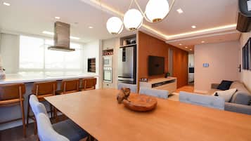 A modern kitchen with a built-in oven, microwave, and refrigerator. There is a dining area with a wooden table and chairs, and a living room with a sofa and a flat-screen TV.