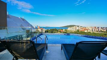 Piscina no terraço com duas espreguiçadeiras, guarda-corpo de vidro e vista para o mar e a paisagem urbana.