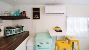 A compact kitchen with a toaster, a small fridge, a white table, and a yellow stool. - Rhonda the Rambler- Glamp w/Pets (Austin)
