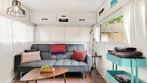 A compact living space with a grey sofa, a wooden coffee table, a teal storage unit, and a window with curtains. - Rhonda the Rambler- Glamp w/Pets (Austin)