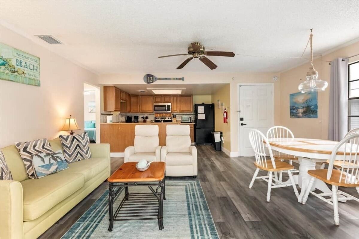 A living room with a sofa, coffee table, and chairs, a dining area with a table and chairs, a kitchen with cabinets and appliances, and a ceiling fan.