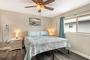 Ein Schlafzimmer mit Bett, Nachttischen, Deckenventilator, Fenster mit Vorhängen und einem Wandbild.