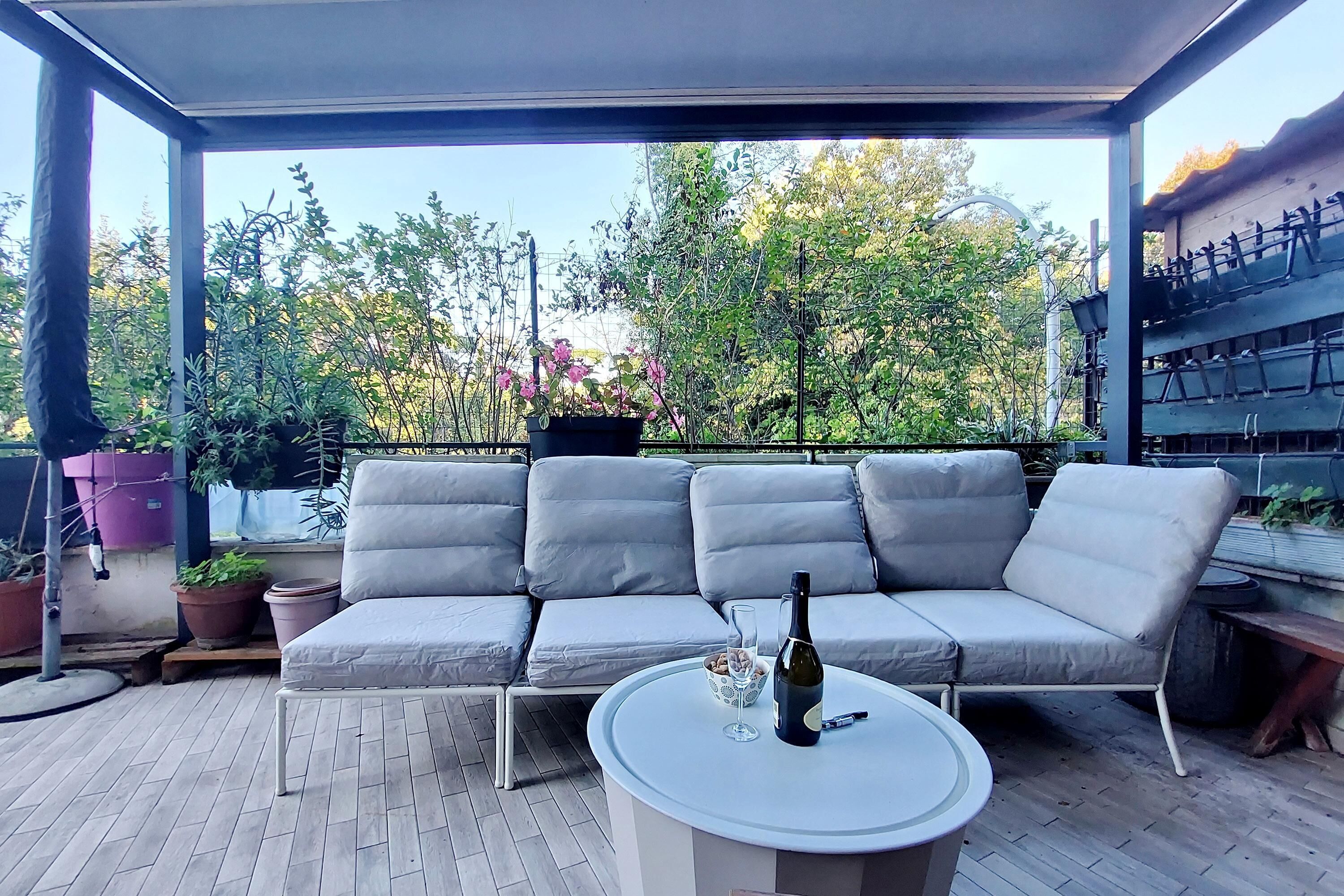 A covered outdoor seating area with a grey sofa and a round table, surrounded by potted plants and a view of trees and buildings.