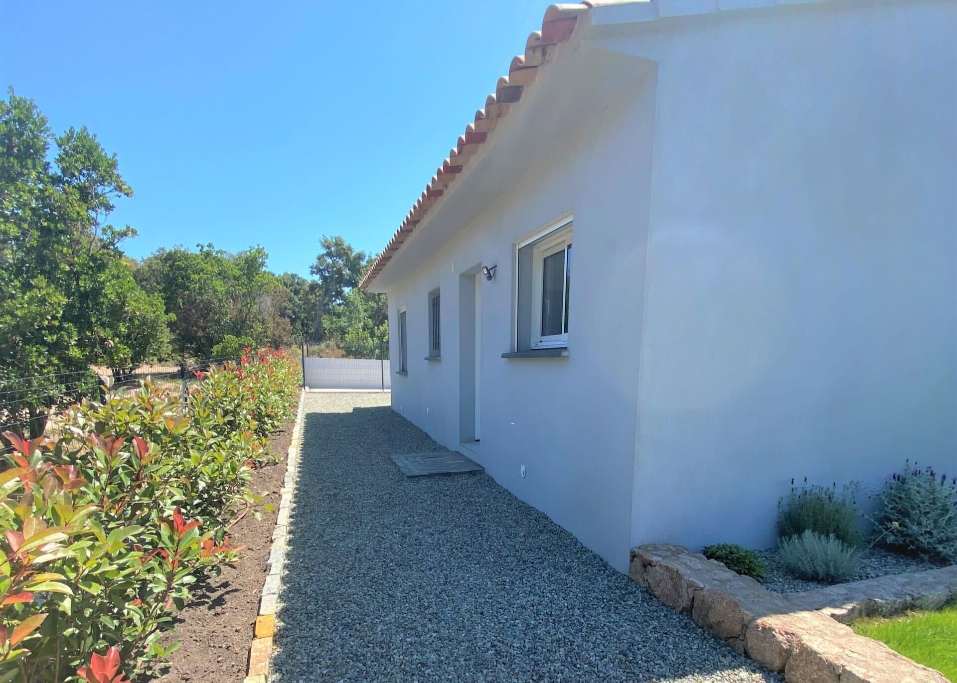 A white building with a red-tiled roof, a gravel driveway, and a garden with red-leafed plants.