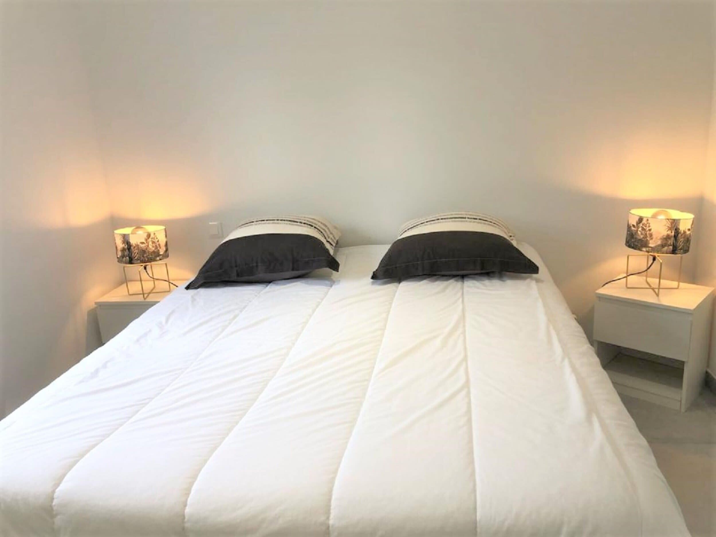 A double bed with white bedding and two black pillows, flanked by nightstands with lamps.