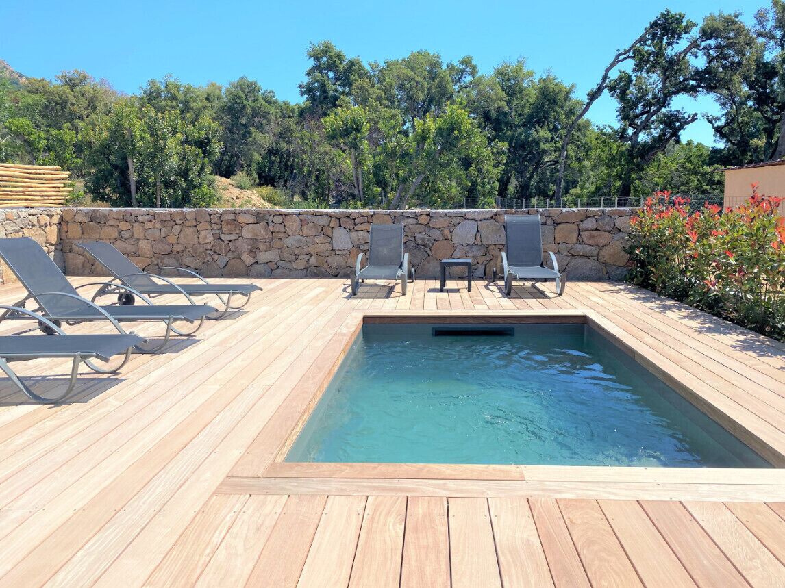 A wooden deck with a rectangular pool, stone wall, and lounge chairs.