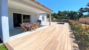 Um deck de madeira com mesa e cadeiras, uma área de varanda coberta e um muro de pedra ao fundo.