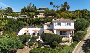 Ein zweistöckiges Haus mit weißer Fassade und einem Ziegeldach, umgeben von üppiger Vegetation und Palmen.