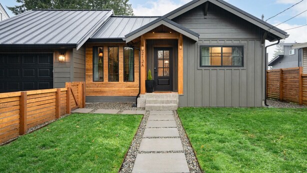 Una casa moderna de dos pisos con fachada gris, un porche de madera y un jardín bien cuidado.