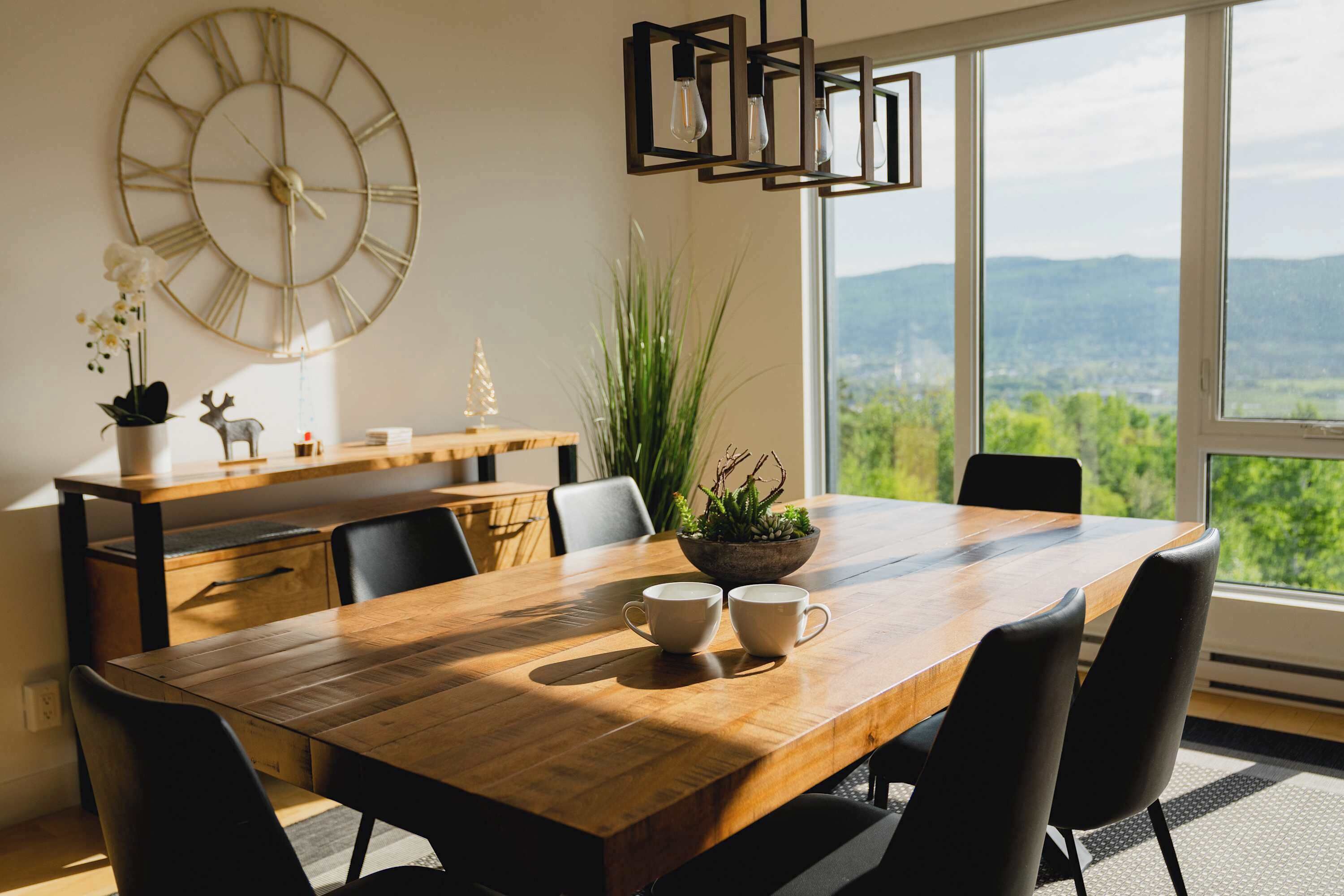 A dining area with a wooden table, black chairs, a large wall clock, and a view of greenery through large windows.