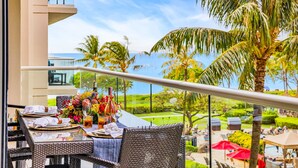 An outdoor dining area with wicker furniture, a table set with plates, glasses, and a floral centerpiece, overlooking a pool and palm trees. - Honua Kai | Ocean View 3 BR, Sleeps 10 | Car Incl. w/ 6+ Nights | HKH ML-2534 by KBM (Lahaina)