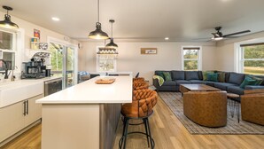 Una cocina moderna con una isla central, taburetes altos, iluminación colgante y un comedor con sofá y mesa de centro.