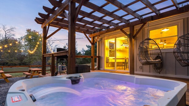 Un jacuzzi bajo una pérgola de madera con luces colgantes, un área de patio y un sillón colgante.
