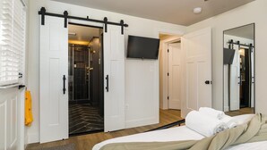 Habitación de hotel con armario corredera, televisor montado, cama con manta color crema y un espejo grande.