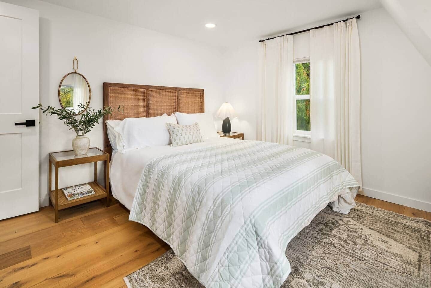 A bedroom with a wooden headboard, a bedside table with a lamp, a window with curtains, and a plant on the nightstand.