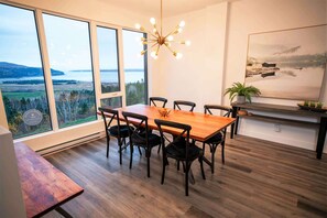 Un espace repas avec une table et des chaises en bois, un lustre et une grande fenêtre donnant sur un lac et des collines.