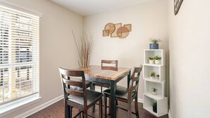 A dining area with a wooden table and chairs, a white shelving unit with plants, and a window with blinds. - Cozy 1Bed 1Bath Apt by Downtown & Farmers Market (Houston)