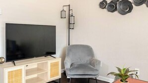 A living room with a flat-screen TV, a grey armchair, a small table with a plant, and a wall-mounted light fixture. - Cozy 1Bed 1Bath Apt by Downtown & Farmers Market (Houston)