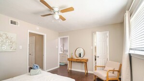 A bedroom with a bed, a chair, a vanity with a mirror, a ceiling fan, and a door leading to a bathroom. - Cozy 1Bed 1Bath Apt by Downtown & Farmers Market (Houston)