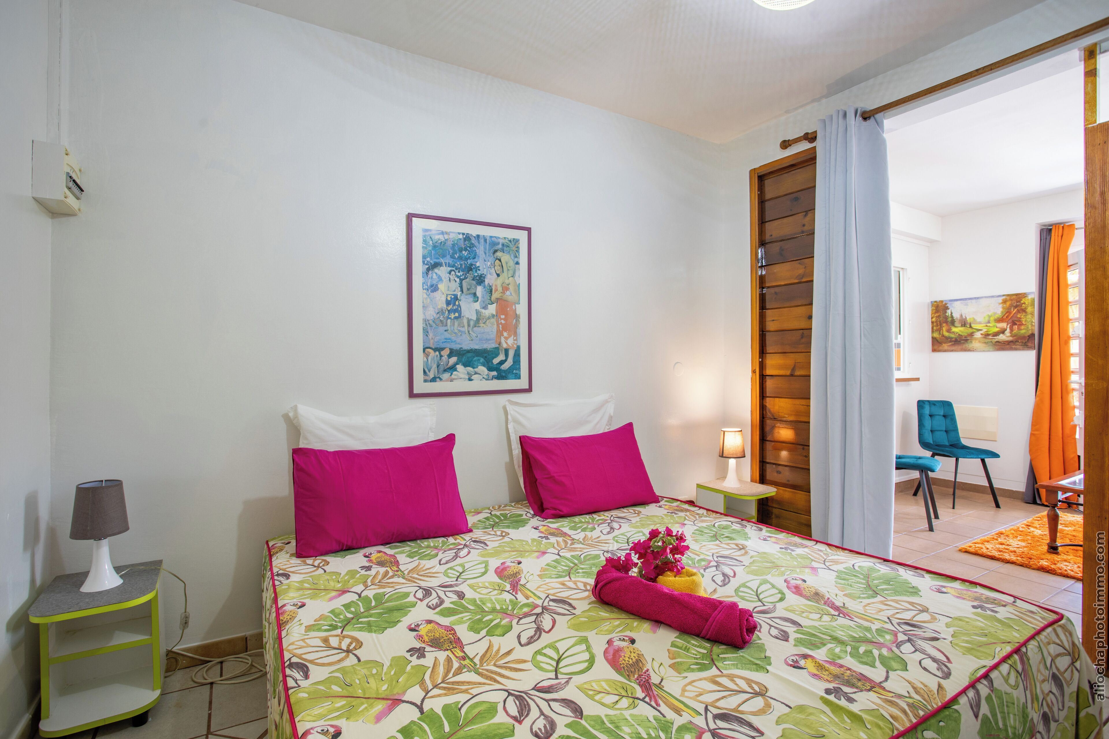 A bedroom with a bed, pillows, a nightstand, a lamp, a painting on the wall, and a view of the adjacent room.