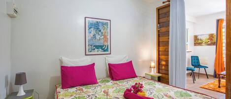 A bedroom with a bed, pillows, a nightstand, a lamp, a painting on the wall, and a view of the adjacent room.