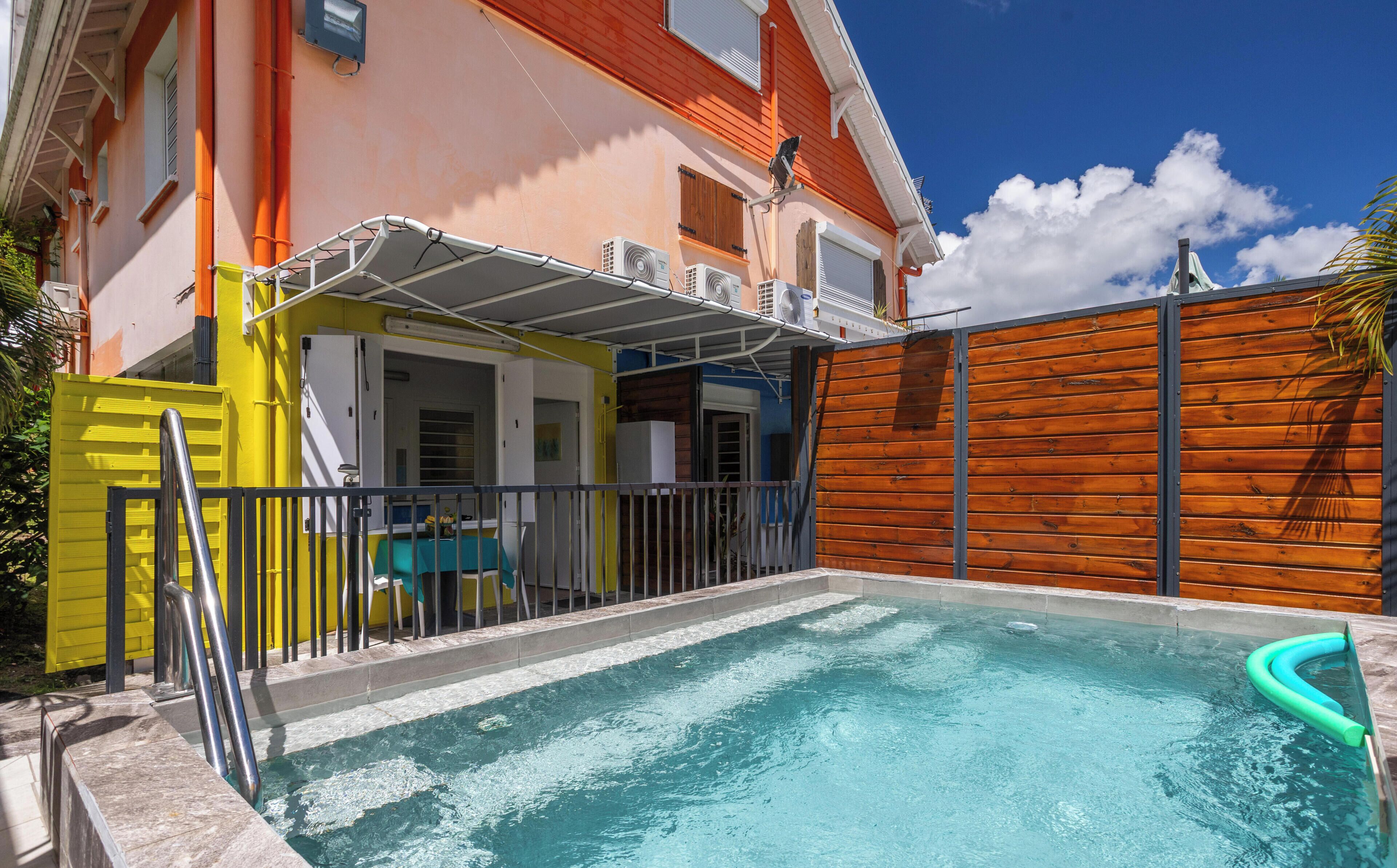 Une piscine entourée d’une clôture en bois, avec une porte jaune et un espace abrité comprenant une table et des chaises.