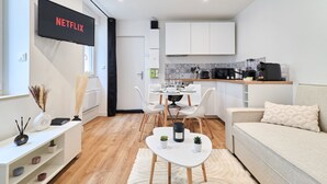 Una cocina moderna con armarios blancos, un comedor con mesa y sillas blancas, y una sala de estar con sofá y televisor mostrando 'NETFLIX'.