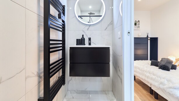 Un baño moderno con tocador negro, espejo redondo con iluminación y una cómoda cama mullida en la habitación contigua.