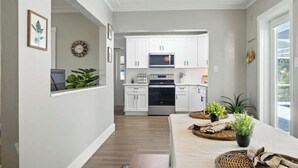 Una cocina moderna con armarios blancos, un microondas y un horno. Hay plantas en el alféizar de la ventana y una zona de comedor con una mesa preparada para dos.