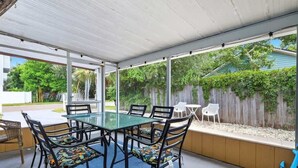 Un patio techado con una mesa y sillas de vidrio, un tapete azul y vistas a una cerca de madera y follaje.