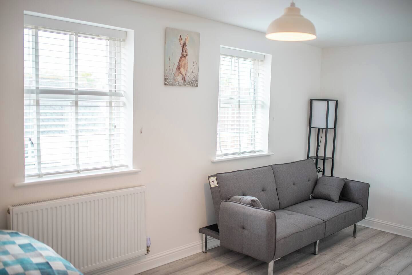 Een moderne woonkamer met een grijze bank, twee ramen met jaloezieën, een radiator en een vloerlamp.