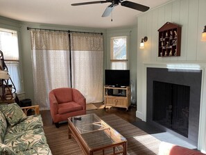 Ein Wohnzimmer mit Sofa, Sessel, Couchtisch, Kamin, Fernseher und dekorativen Regalen.