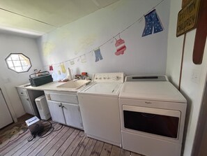 Ein Waschraum mit Waschmaschine, Trockner und Waschbecken. Ein Fenster mit sechseckigem Muster und eine Wäscheleine mit verschiedenen Kleidungsstücken.