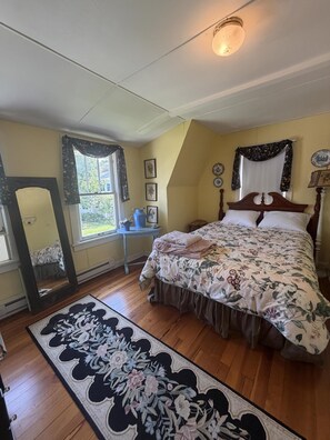 Ein Schlafzimmer mit einem großen Bett, einem fensterbelaubten Fenster, einem Spiegel und einem kleinen Tisch mit einer Vase.