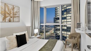 A modern hotel room with a large window offering a city view, a bed with white linens, a nightstand with a lamp, and a chair. - Radiant Boulevard by DVH Luxe Retreats (Abu Dhabi)