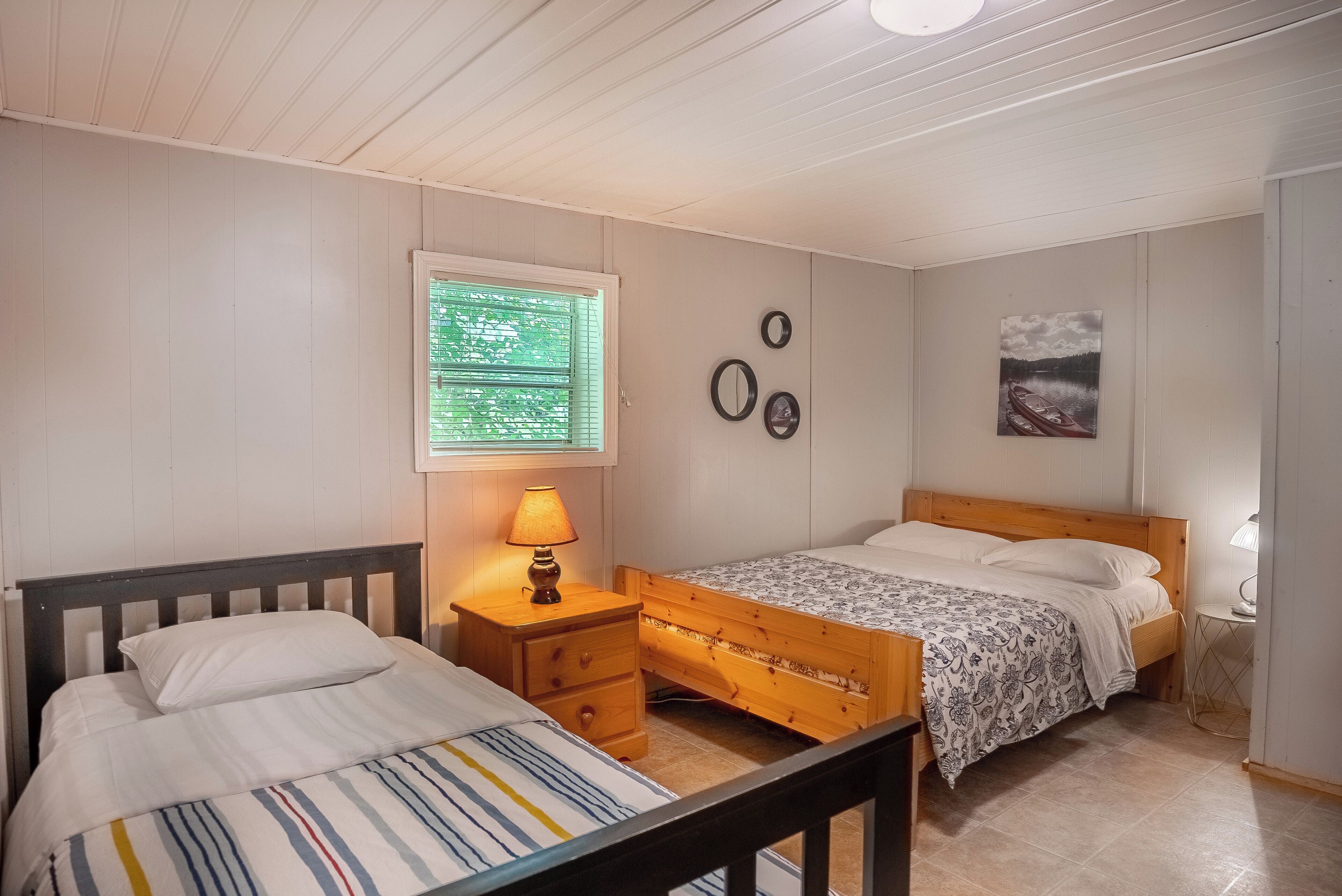 A bedroom with two beds, a window with blinds, a bedside table with a lamp, and wall decorations.