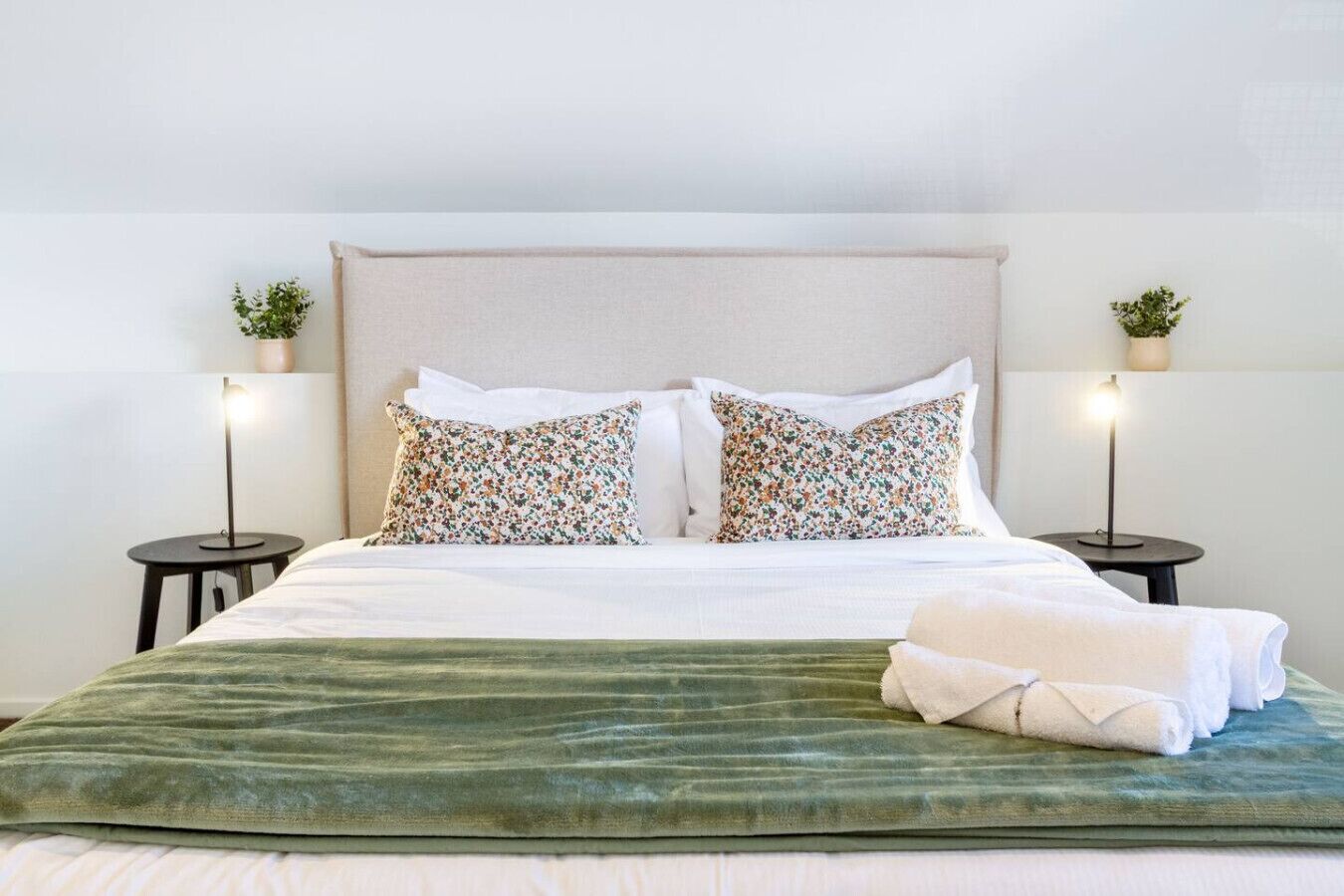 A neatly made bed with a green bedspread and two floral pillows, flanked by two small tables with lamps and potted plants.
