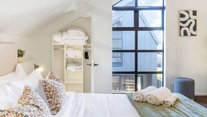 Un dormitorio con un ventanal grande, una cama con almohadones florales, un armario con toallas dobladas y un otoman gris.