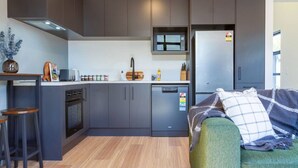 Cocina compacta con armarios oscuros, microondas, lavavajillas y refrigerador. Se observa un pequeño comedor con taburetes y una mesa. En primer plano, un sofá verde con una manta escocesa.