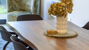 Ein Essbereich mit einem Holztisch, Stühlen, einer Vase mit getrockneten Blumen und einem an der Wand befestigten Fernseher.