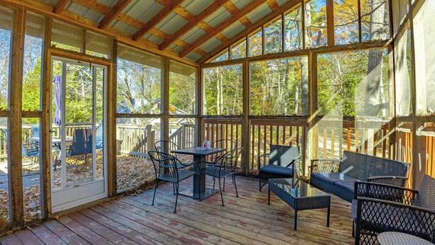Une terrasse extérieure couverte, dotée d’un plancher en bois, de mobilier en métal et de grandes fenêtres offrant une vue sur les arbres.