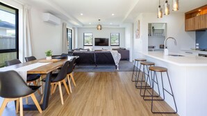 A modern kitchen with a central island, bar stools, and a dining area with a table and chairs. - Halswell Quarry - Walk and Explore - 3 bed (Christchurch)