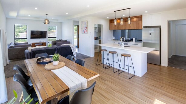 A modern kitchen with a central island, bar stools, and a dining area with a wooden table and chairs. - Halswell Quarry - Walk and Explore - 3 bed (Christchurch)