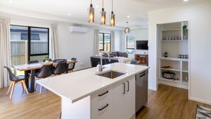 A modern kitchen with a central island, a dining area with a table and chairs, and a built-in bookshelf. - Halswell Quarry - Walk and Explore - 3 bed (Christchurch)