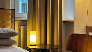 Een hotelkamer met een bed, nachtkastje met lamp, stoel en raam met gordijnen.